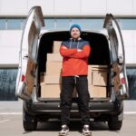 Man in red jacket and blue beanie stands by van of boxes in the city, ready for delivery.
